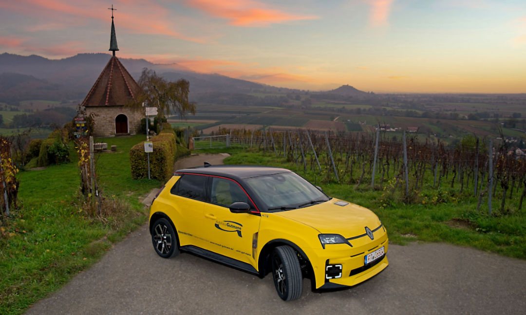 Ein gelber Renault 5 mit schwarzem Dach steht auf einem schmalen Weg inmitten einer idyllischen Weinberglandschaft bei Sonnenuntergang. Im Hintergrund ist eine kleine Kapelle mit spitzem Turm zu sehen, daneben ein Wegweiser. Der Himmel ist in warmen Farben gefärbt, mit Orangetönen und rosa Wolken. Die hügelige Landschaft erstreckt sich bis zum Horizont.