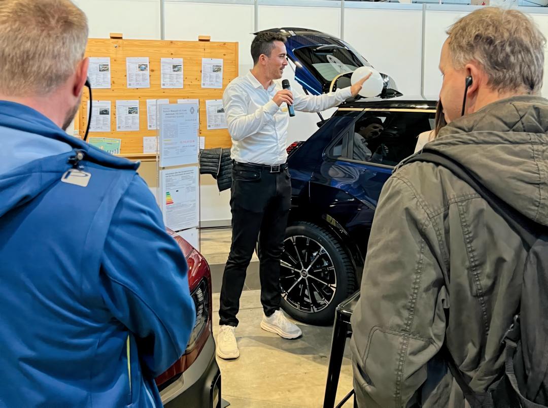 Luca Gutmann erklärt Besuchern ein ausgestelltes Fahrzeug mit geöffnetem Kofferraum während einer Präsentation auf der Messe.