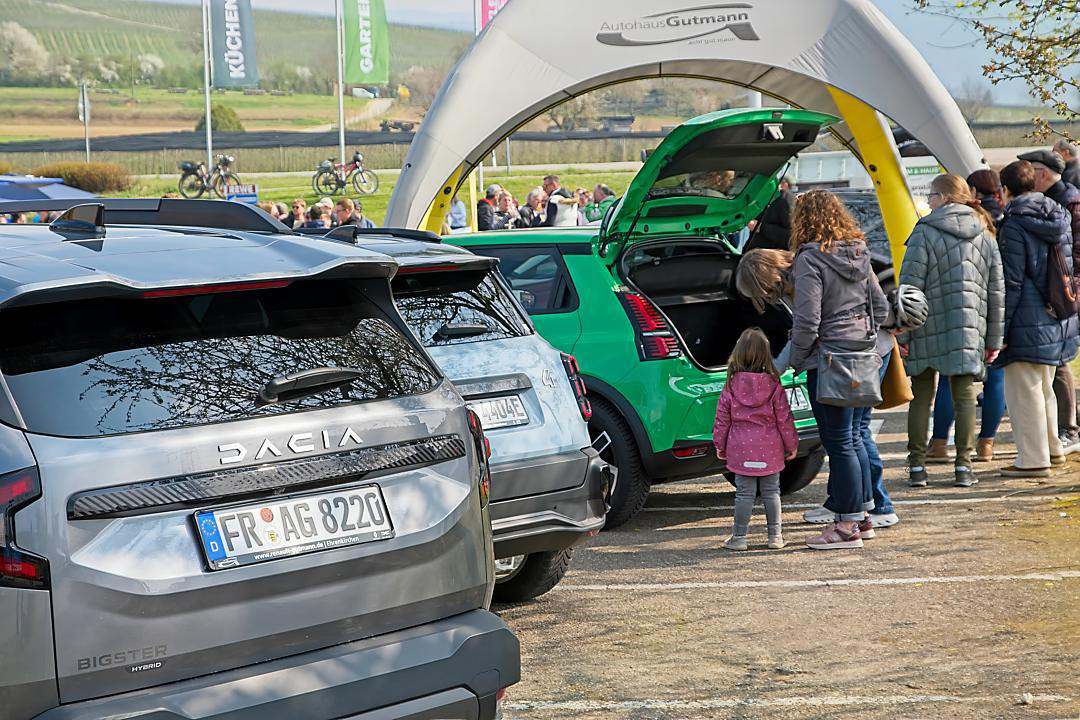 Eine Familie mit einem kleinen Mädchen schaut sich einen grünen R5 E-tech mit geöffneten Türen an. Das Kind zeigt auf das Auto, während die Eltern mit einer Verkäuferin sprechen. Daneben steht ein grauer Dacia Bigster Hybrid.
